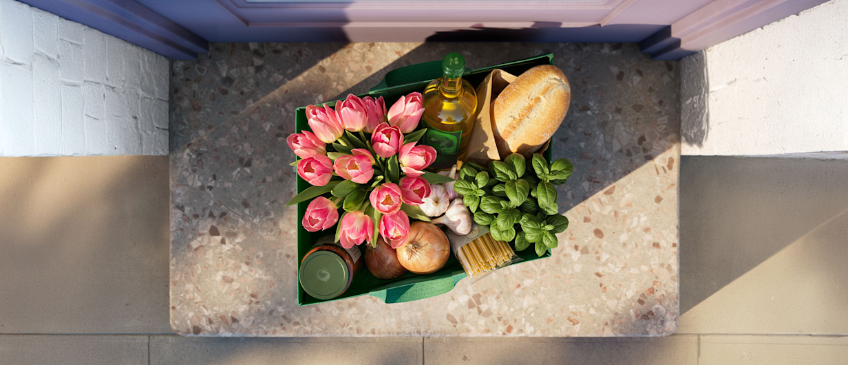 Overhead view of a door stoop with a grocery delivery bag containing flowers, bread, olive oil and produce, where the bag's shape forms SynaPay's parallelogram logo.